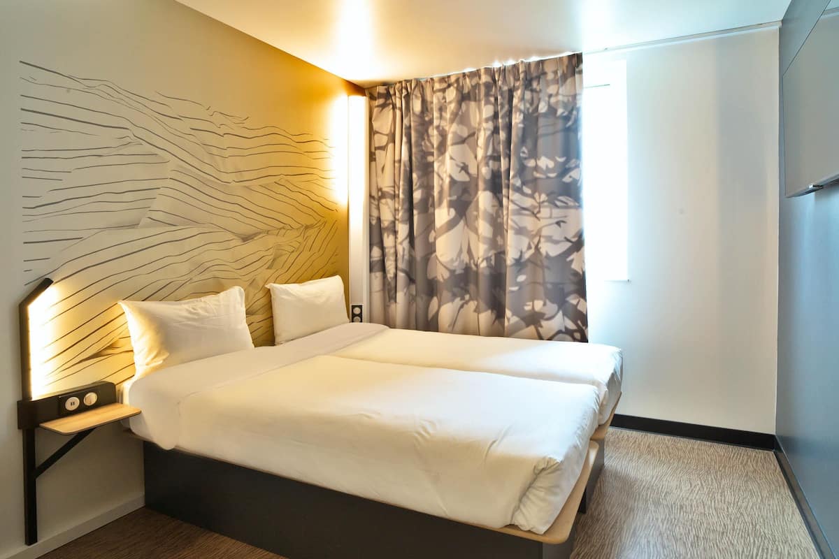 A double bed with white linens, a nightstand with a lamp, and a wall with a leaf pattern.