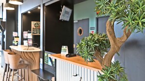A reception area with a wooden counter, a digital screen, and a potted tree. - B&B HOTEL Dreux Nord (Dreux)
