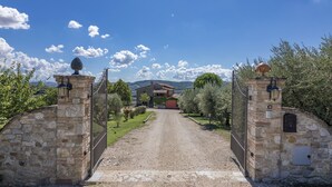 A stone gate leading to a rustic house with a red roof, surrounded by greenery and mountains in the background. - Villa Le Logge 12, Emma Villas (Todi)