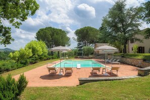 A rectangular pool with a tiled deck, surrounded by lounge chairs and umbrellas, set against a backdrop of trees and a building.
