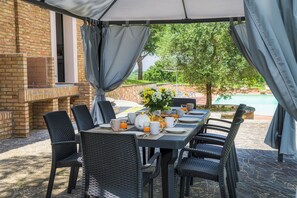 An outdoor dining area with a brick wall, a grill, and a pool in the background.