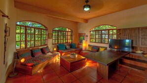 Una sala de estar acogedora con un conjunto de sofá, una mesa de centro y una televisión. Amplias ventanas con arcos superiores dejan entrar luz natural y ofrecen vistas al exterior con vegetación.