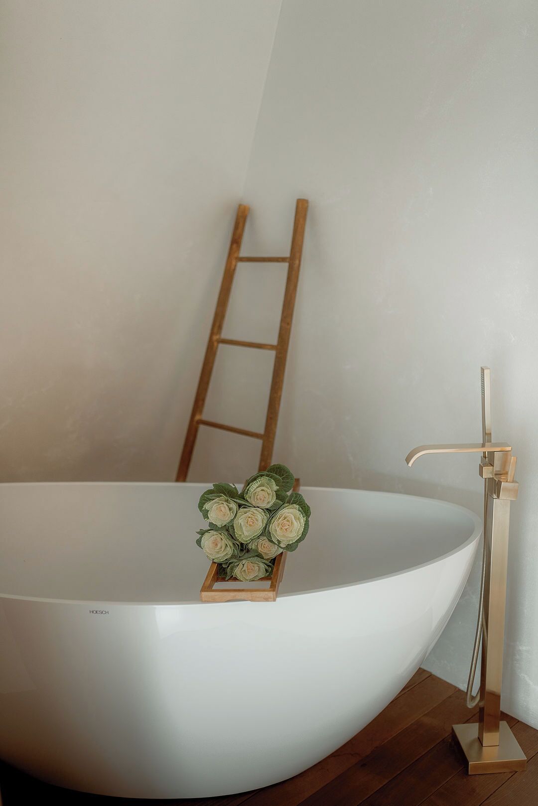 Una bañera grande y blanca con una escalera de madera apoyada en la pared, una pequeña caja de madera con flores artificiales y un grifo dorado.