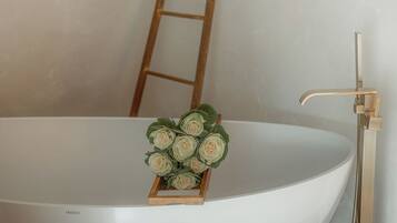 Una bañera grande y blanca con una escalera de madera apoyada en la pared, una pequeña caja de madera con flores artificiales y un grifo dorado.