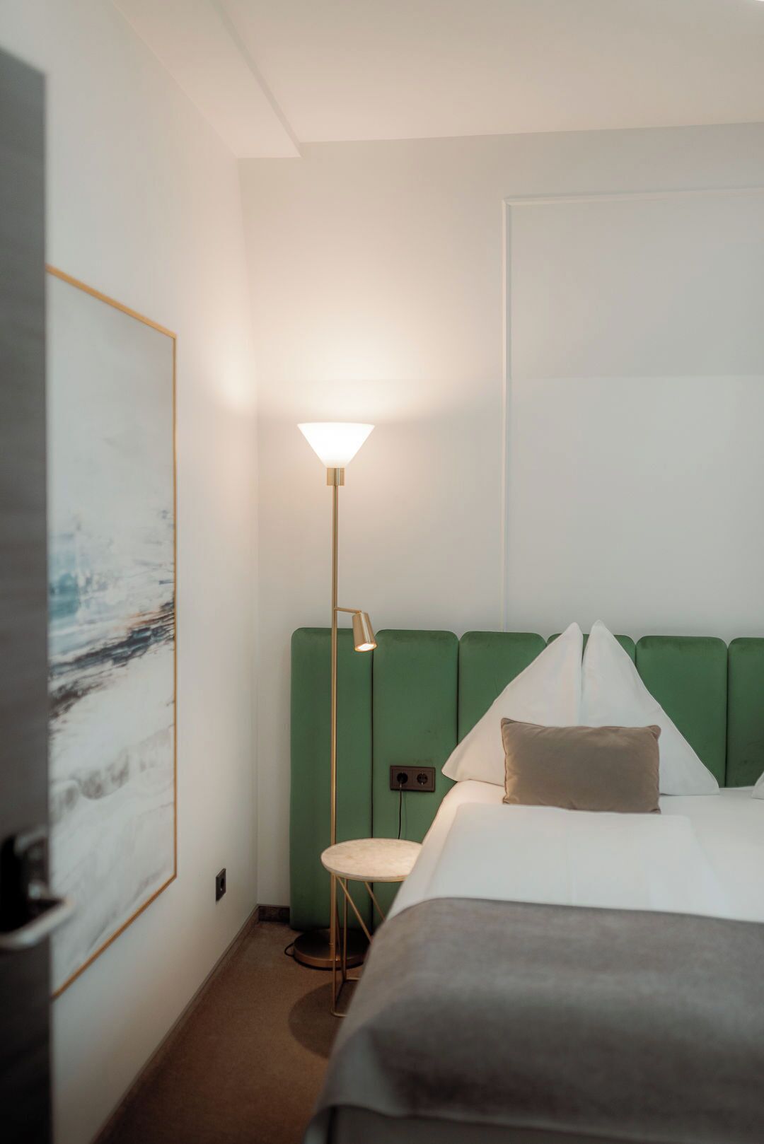 A hotel room with a bed, a green headboard, a floor lamp, and a painting on the wall.