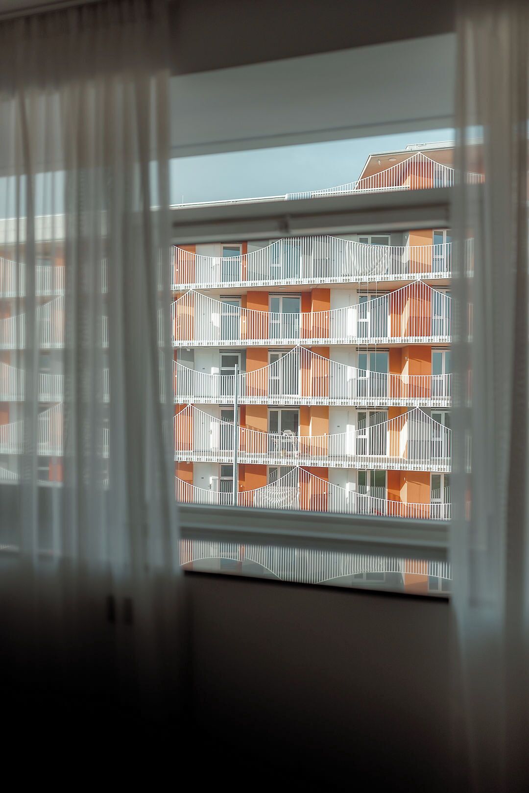 Vista a través de una ventana con persianas blancas, mostrando un edificio de varios pisos con balcones y detalles color naranja.