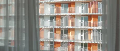 Vista a través de una ventana con persianas blancas, mostrando un edificio de varios pisos con balcones y detalles color naranja.