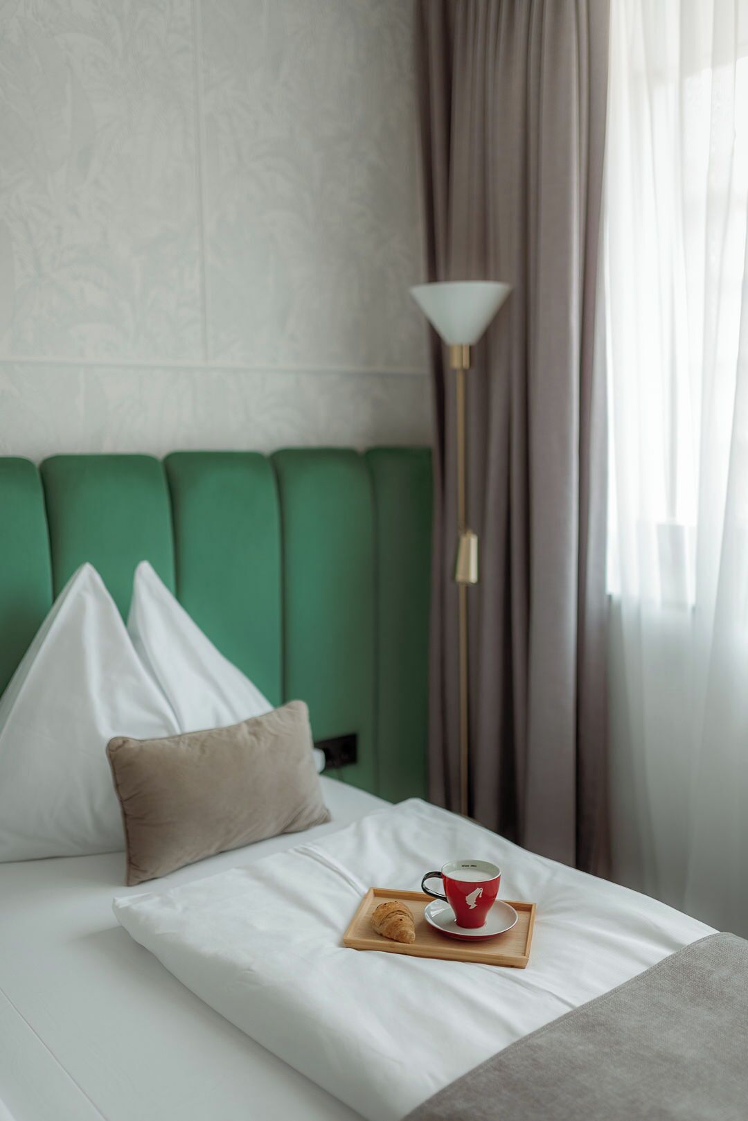 A neatly made bed with a green headboard, a bedside lamp, and a tray with a cup and pastry.