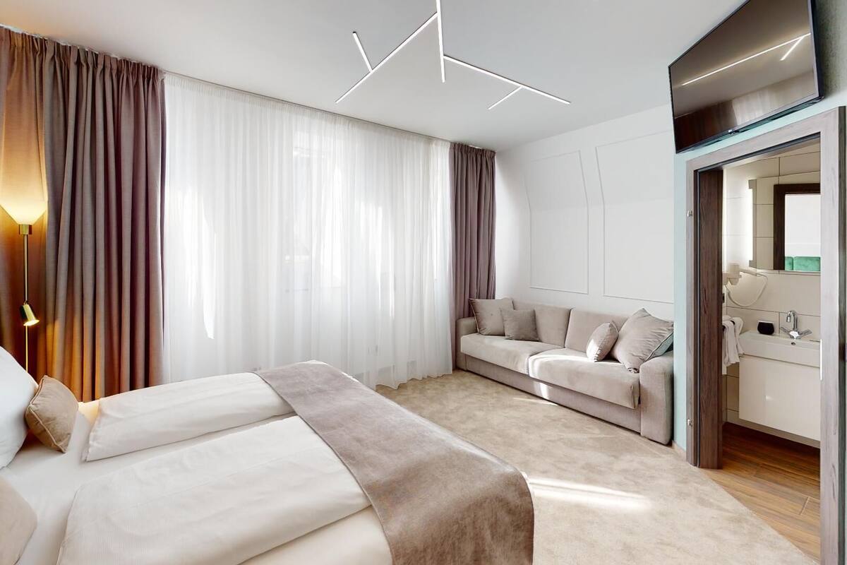 Una habitación de hotel moderna con una cama, un sofá, un espejo y un baño visible a través de una puerta abierta.