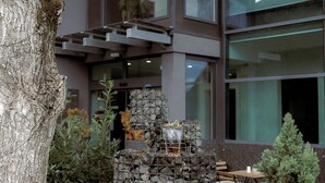 Un bâtiment moderne agrémenté d’une fontaine en pierre, d’un grand arbre et d’un espace de détente avec des tables et des chaises.