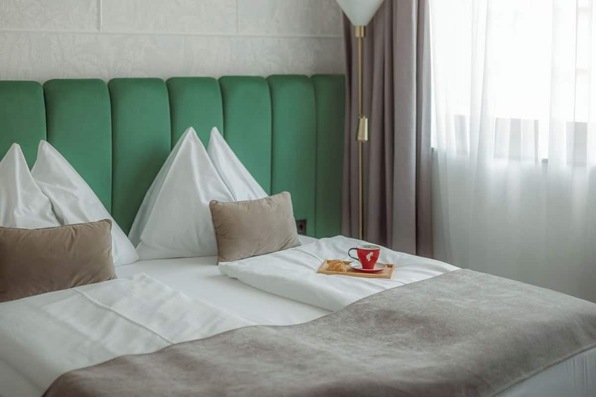 Habitación de hotel con cabecera tapizada en verde, ropa de cama blanca y una mesita de noche con una taza y un pastel.