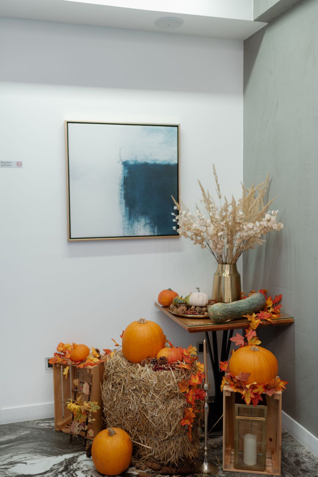 Un coin de pièce avec un tableau abstrait encadré, une petite table décorée sur le thème de l’automne et un vase contenant des plantes séchées.