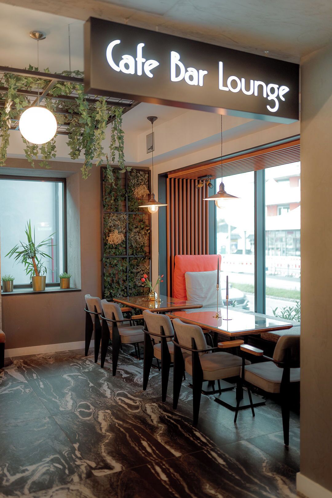 A cafe bar lounge with a modern interior, including a wooden table, chairs, and a large window offering a view outside.