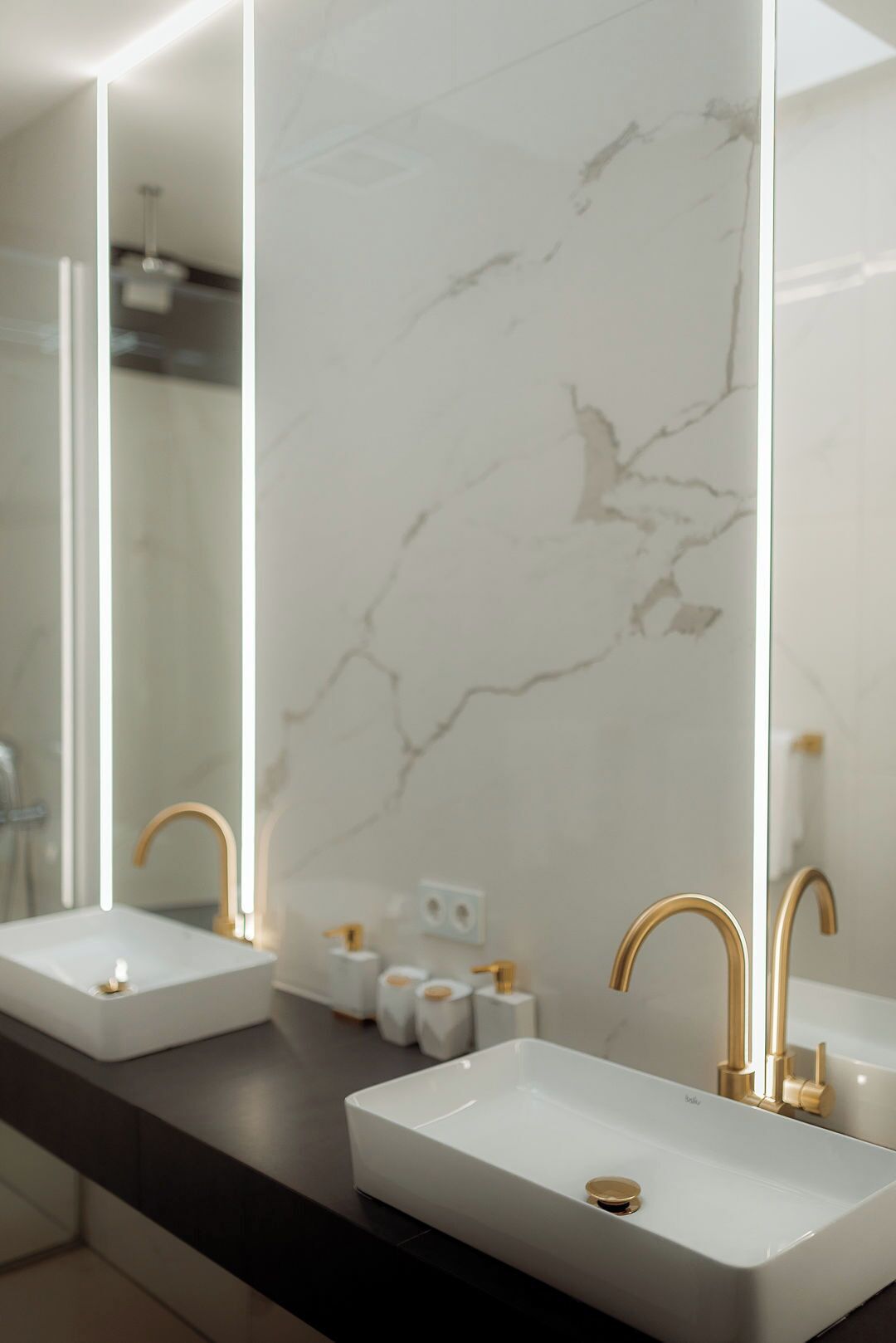A bathroom with two white sinks, gold faucets, and a marble backsplash.