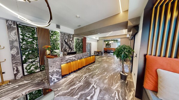 A modern hotel lobby with a marble reception desk, indoor plants, and a seating area with orange cushions. - NED Hotel (Graz)