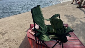 A beach with a green folding chair and a colorful mat on the sand, overlooking a calm sea with boats and distant hills. - Ayci Otel (Bodrum)