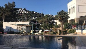 A swimming pool with lounge chairs, a small building, and a hill with houses in the background. - Ayci Otel (Bodrum)