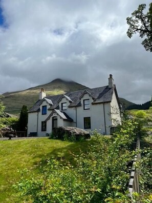 Traditional two-story house with a steeply pitched roof, set amidst a lush landscape with a well-manicured lawn and surrounding trees.