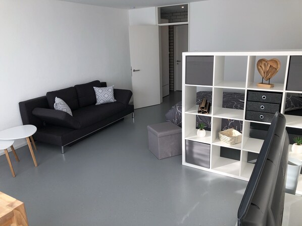 A modern living room with a black sofa, a white coffee table, and a grey ottoman. There is a white shelving unit with a TV on top. - Great apartment with style and a view of Talheim (Talheim)