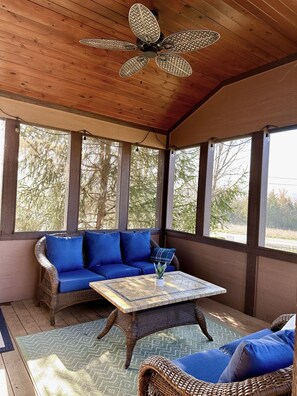 A covered porch with a ceiling fan, wicker furniture, and large windows offering views of trees.