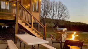 Una terrassa de fusta amb taula i caires de picnic, un parell de caires de barbacoa amb foc, i una escala que condueix a un edifici amb llums de garlandes.