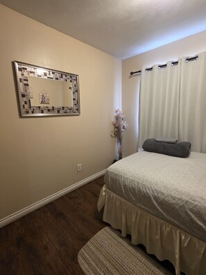 A bedroom with a bed, a mirror, a window with curtains, a vase with flowers, and a rug. - Last frontier Rental Family #3 (Anchorage)