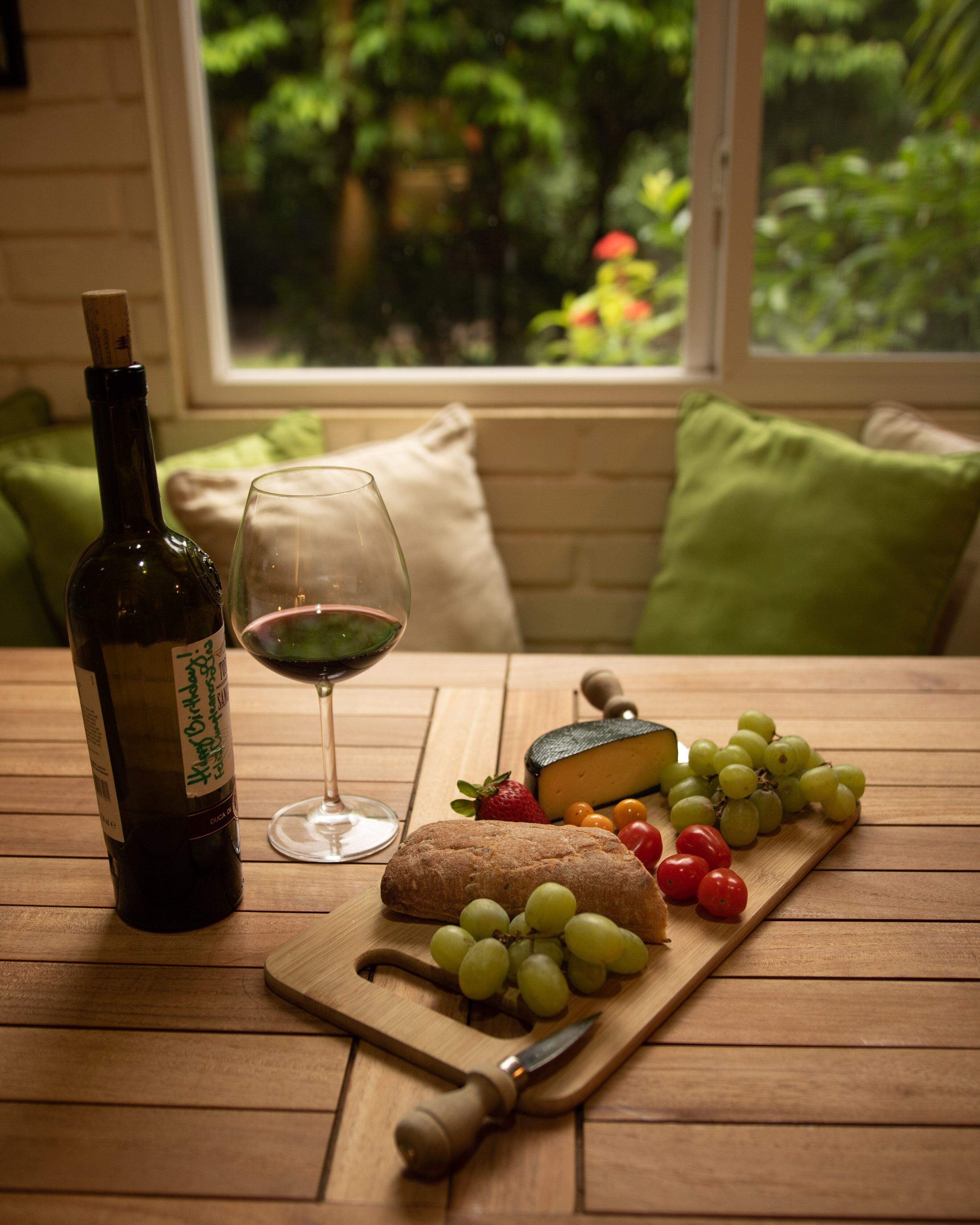 A wooden table with a wine bottle, a glass of red wine, a loaf of bread, a cheese, and a variety of grapes and tomatoes.