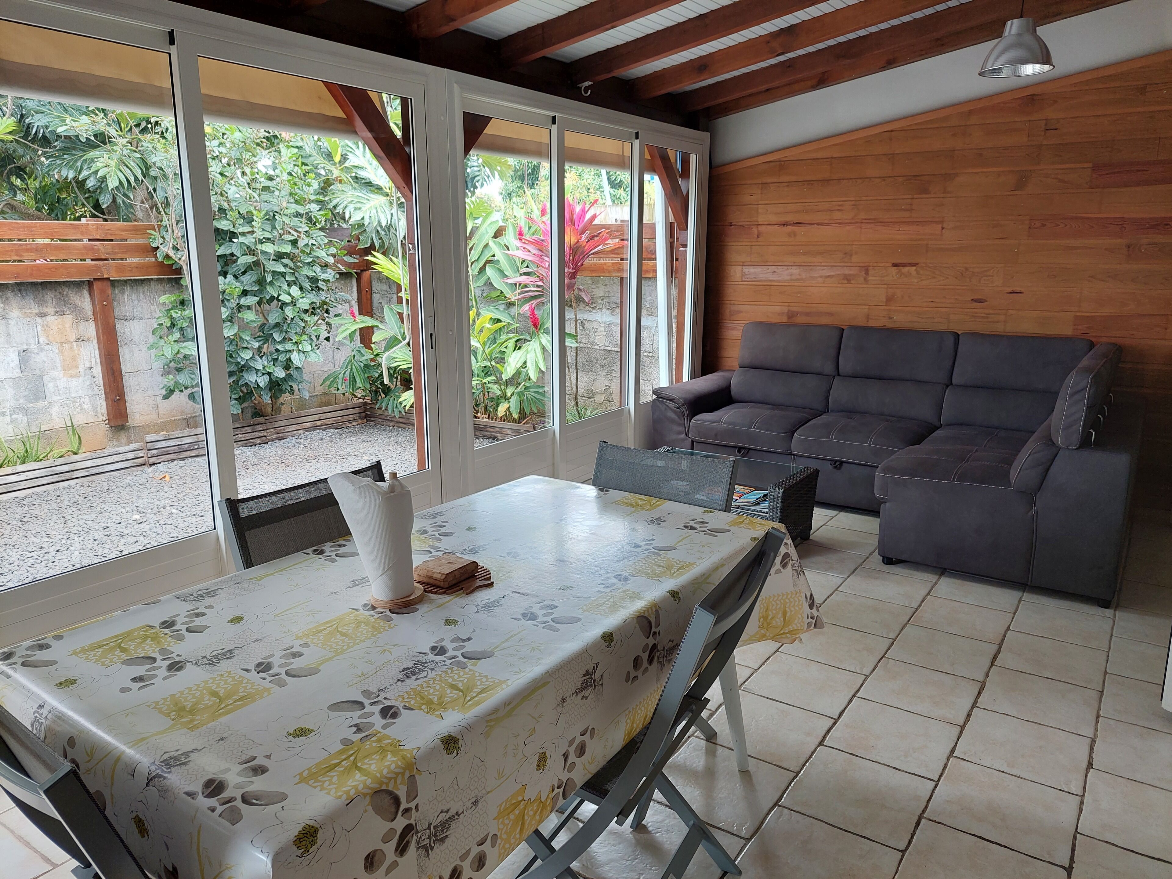 A cozy room with a sofa, a table with a floral tablecloth, and large windows offering a view of greenery outside.