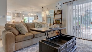 A modern living room with a sofa, a coffee table, and a TV stand. There is a kitchen in the background with a large window and a clock on the wall. - Safety Harbor Paradise (Safety Harbor)