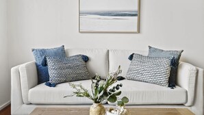A white sofa with patterned cushions, a framed picture on the wall, and a vase with plants on the floor.