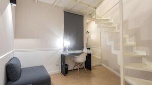 A modern room with a desk, chair, and a small sofa. There is a staircase with a white railing and a window with blinds. - Five Boutique Rooms - Camera 1 (Naples)