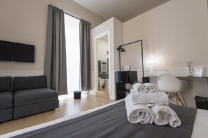 A modern hotel room with a grey sofa, a neatly made bed with rolled towels, a flat-screen TV, a desk with a lamp, and a view of the bathroom. - Five Boutique Rooms - Camera 5 (Naples)