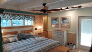 Een slaapkamer met een groot bed, een houten hoofdbord, een plafondventilator, een raam met bloemen gordijnen, een bedlamp en een deur met uitzicht.