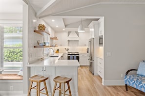 Une cuisine moderne avec des meubles blancs, un îlot central doté de tabourets et une vue sur l’extérieur grâce à de grandes fenêtres.