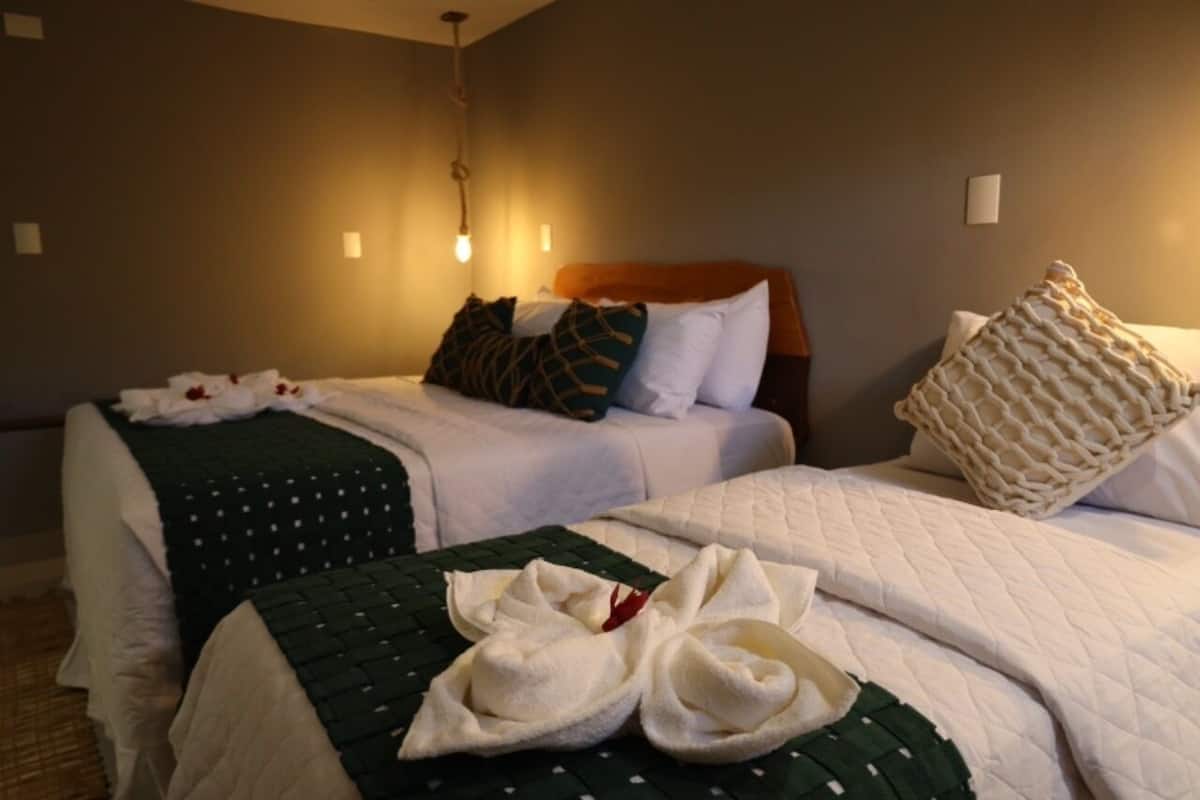 Quarto de hotel com duas camas, cada uma com colcha verde e branca. Há várias almofadas, um cobertor de tricô e um par de toalhas brancas nas camas.