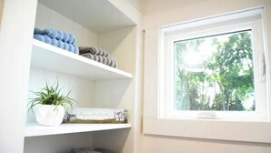 Une salle de bain avec des étagères blanches, une plante en pot et une fenêtre donnant sur des arbres.