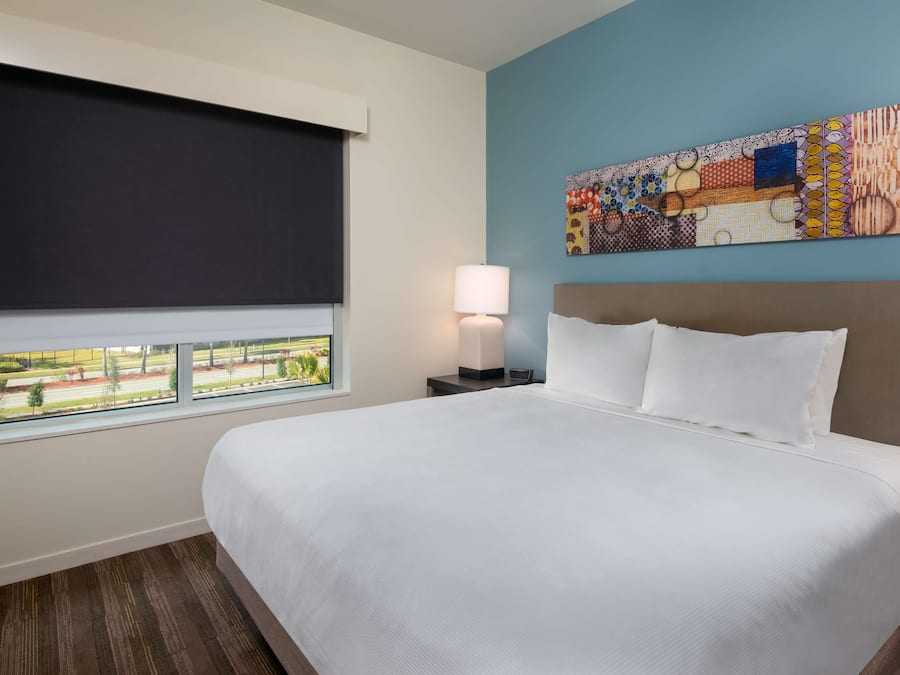 Ein Hotelzimmer mit einem großen Bett, einem Fenster mit Blick auf eine angelegte Grünfläche, einem Nachttisch mit Lampe und einem farbenfrohen, abstrakten Gemälde an der Wand.