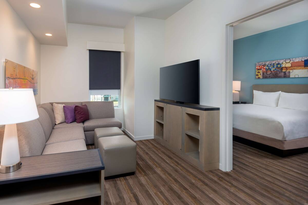 A modern hotel room with a sofa, ottoman, flat-screen TV, and a bed.