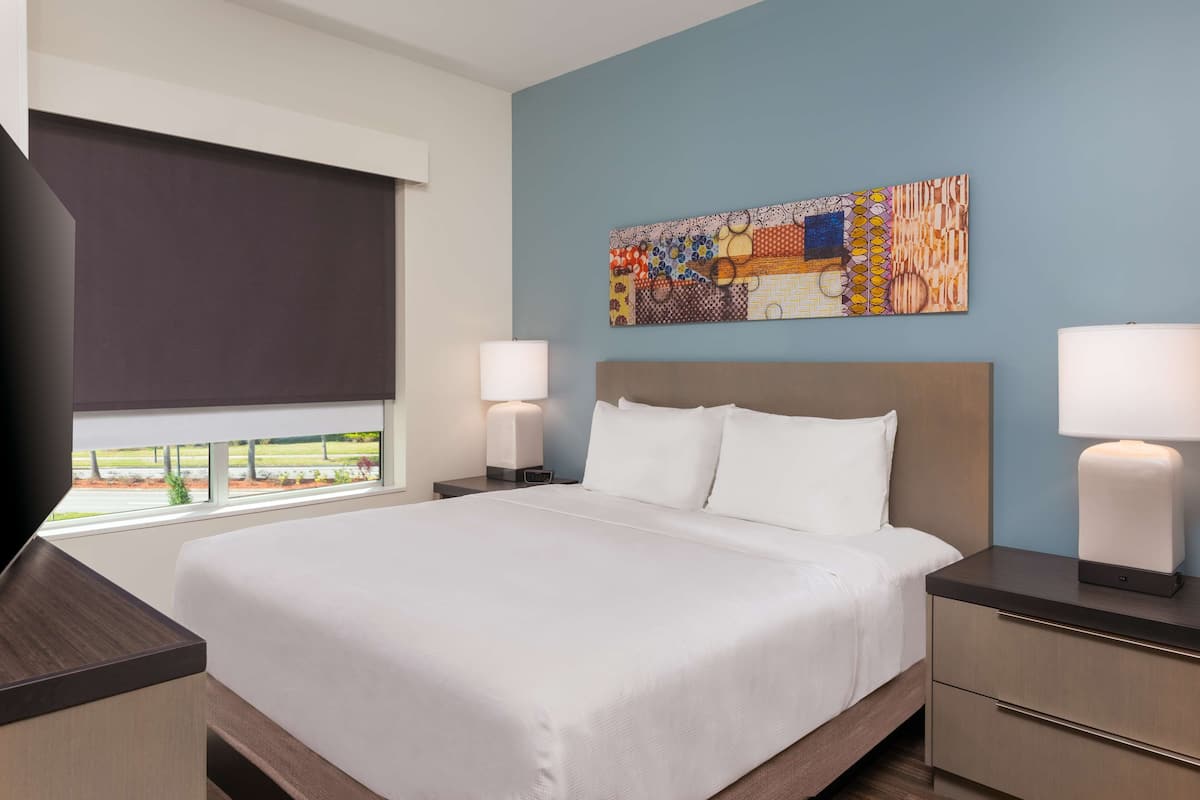 A hotel room with a large bed, two bedside tables with lamps, a window with a view, and a colorful abstract painting on the wall.