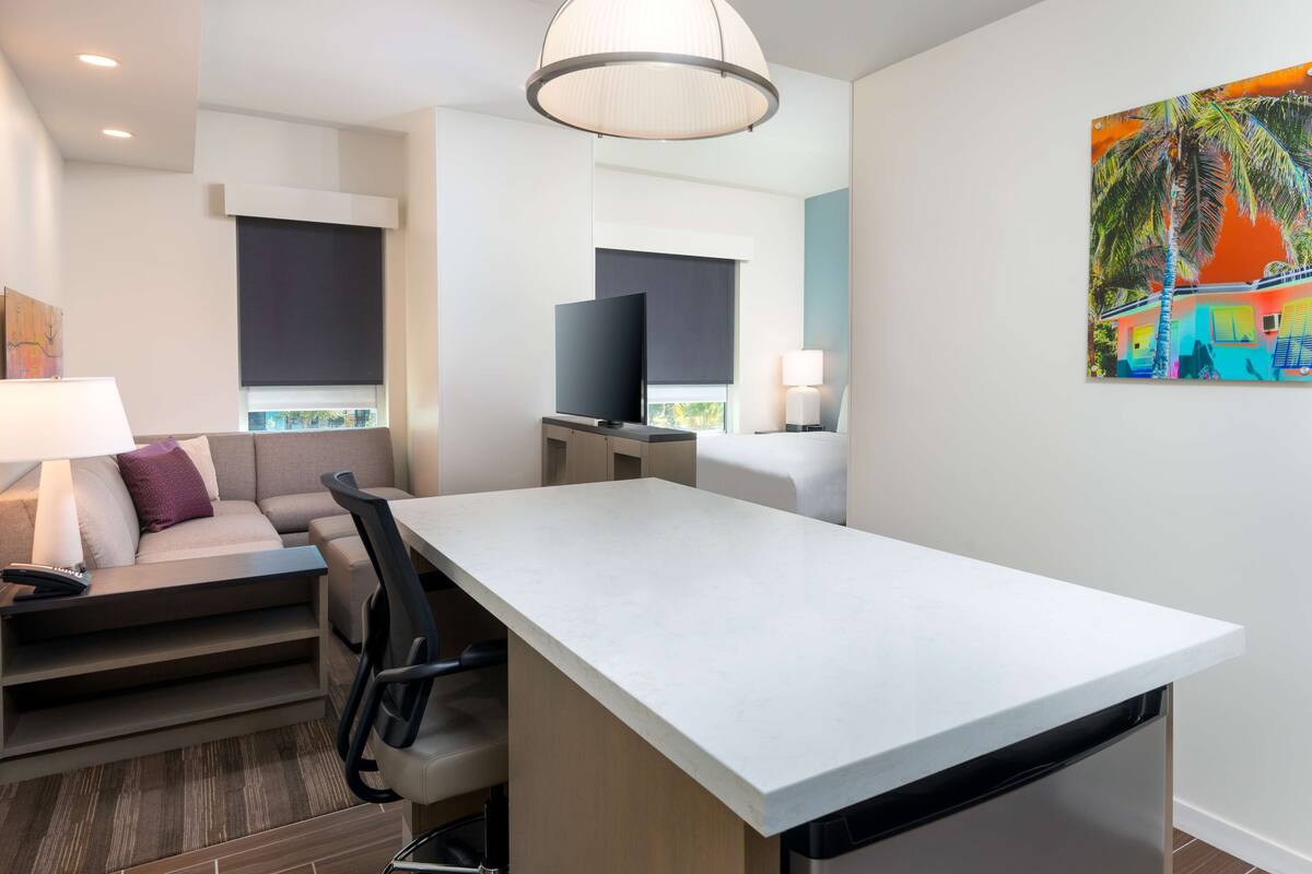 A modern hotel room with a dining area, a sofa, a TV, and a painting on the wall.