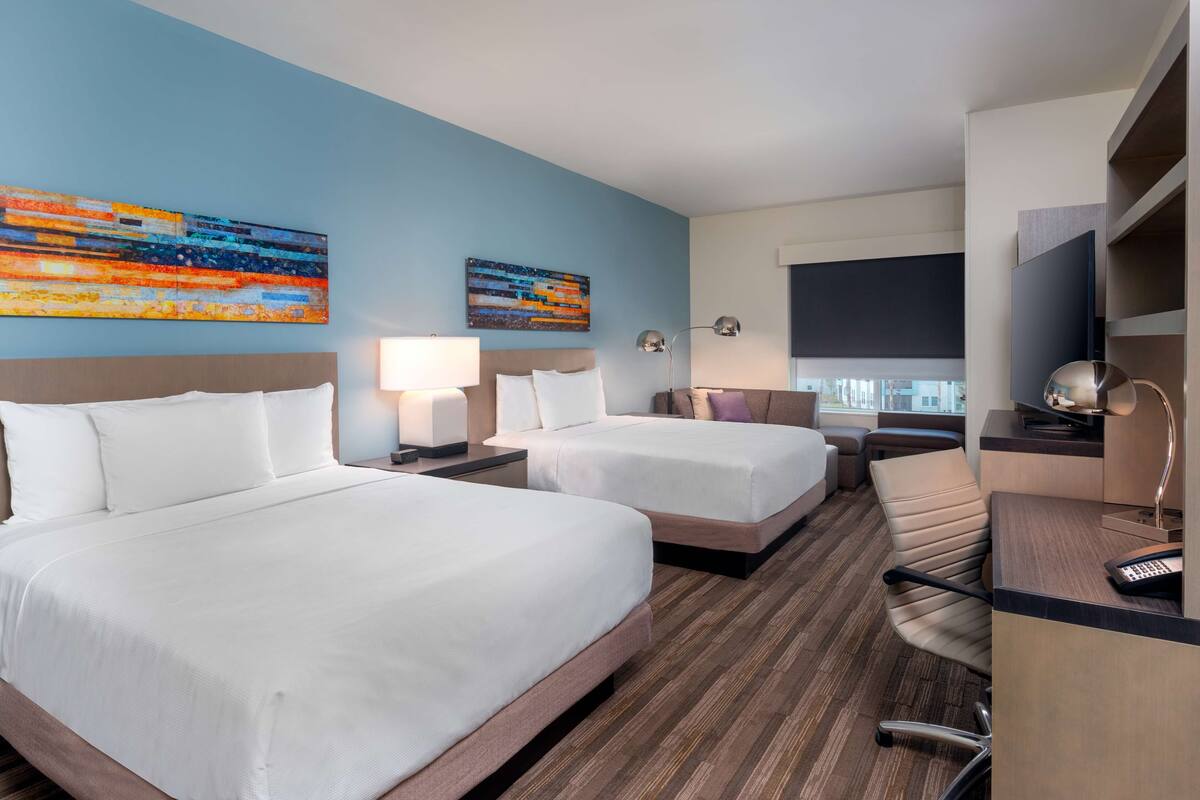 A hotel room with two beds, a desk, a chair, a lamp, and two paintings on the wall.