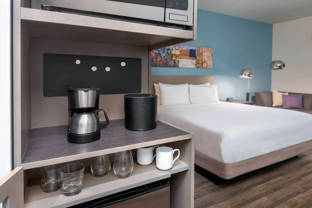 A hotel room with a bed, a coffee maker, a microwave, and a shelf with glasses and mugs.