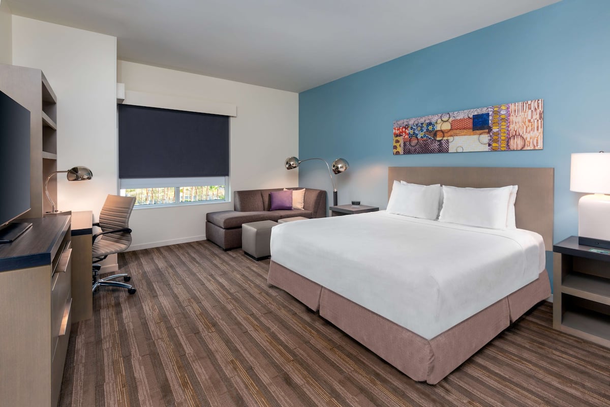 A hotel room with a large bed, a desk, a chair, a sofa, a TV, and a window with blinds.