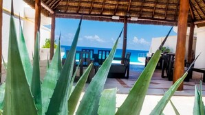 A beachside patio with a view of the ocean, featuring a thatched roof, outdoor seating, and large green plants. - Beautiful 2 levels apartment at the best beach area of Cancun hotel zone.... (Cancún)