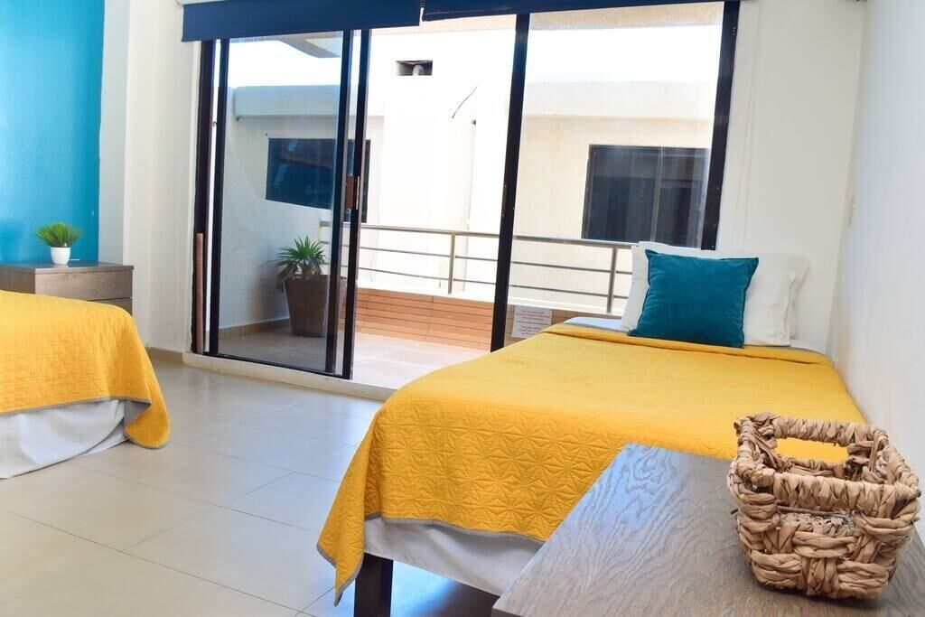 A hotel room with a bed, a small table, a wicker basket, and a balcony with a railing.