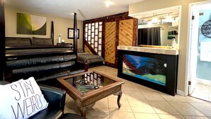 Una sala de estar con un sofá de cuero negro, una mesa de centro de vidrio, una chimenea con protector decorativo y una escalera con paneles de madera.