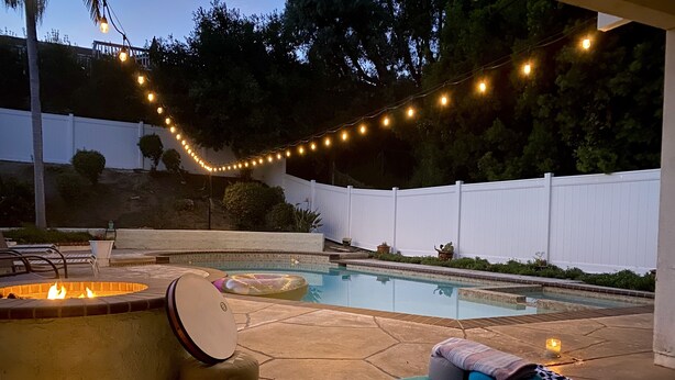 Un patio trasero con piscina, fogata y luces colgantes.