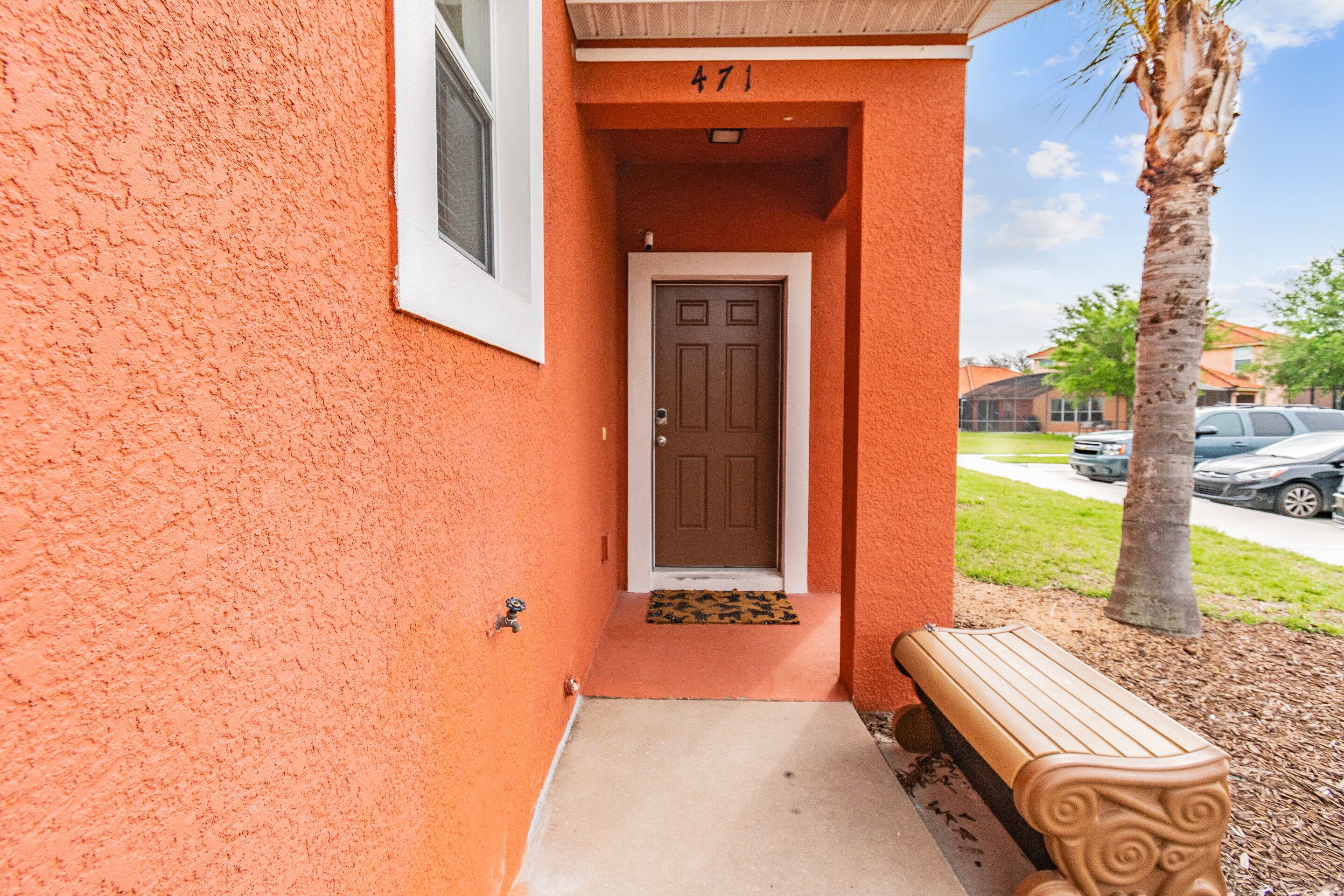 A building entrance with a brown door, a number '471' above it, a small bench outside, and a palm tree nearby.