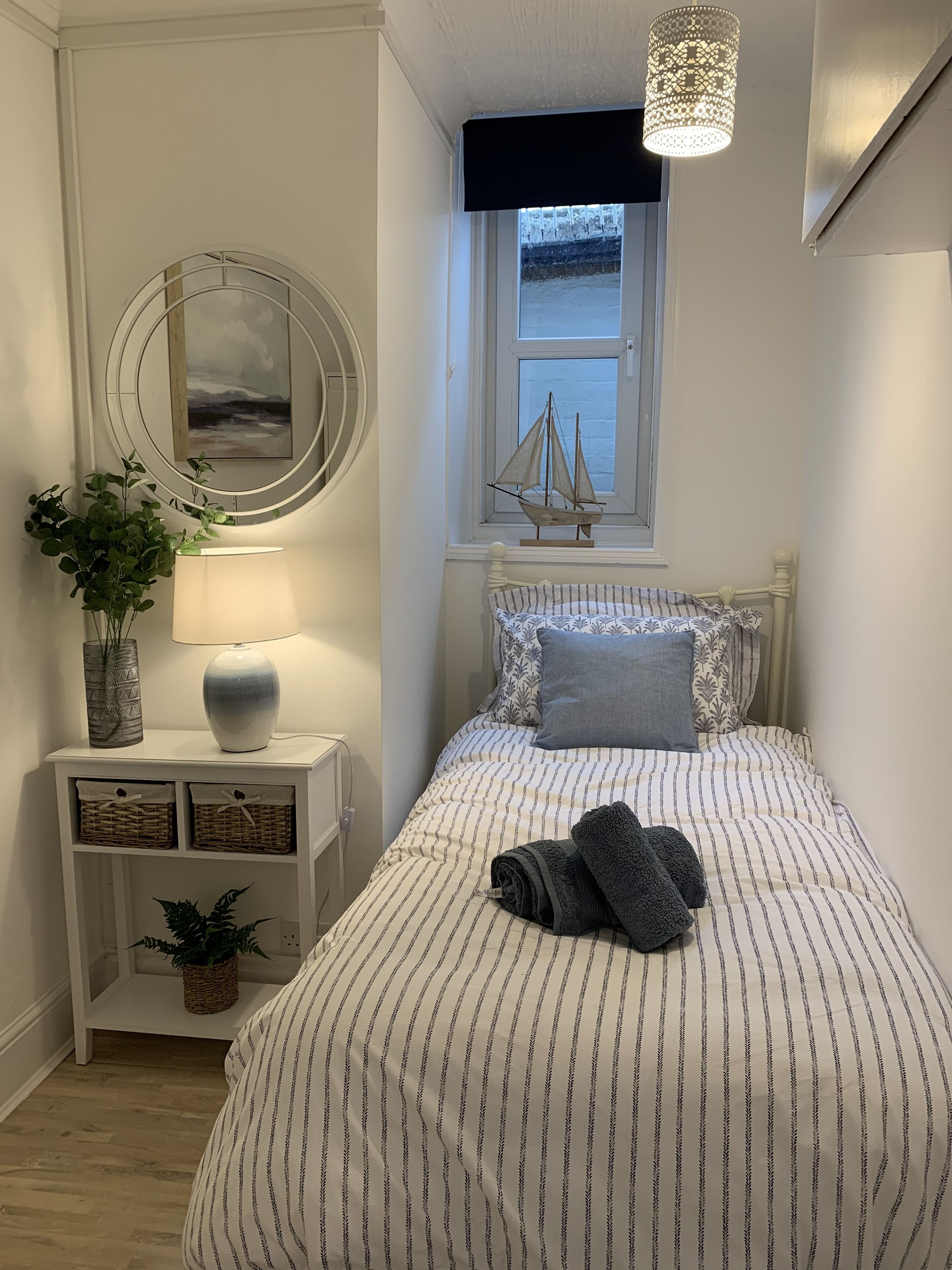 A neatly arranged bedroom with a bed, a bedside table with a lamp, a decorative mirror, and a window with a sailboat model.
