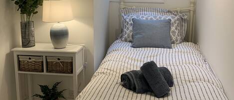 A neatly arranged bedroom with a bed, a bedside table with a lamp, a decorative mirror, and a window with a sailboat model.